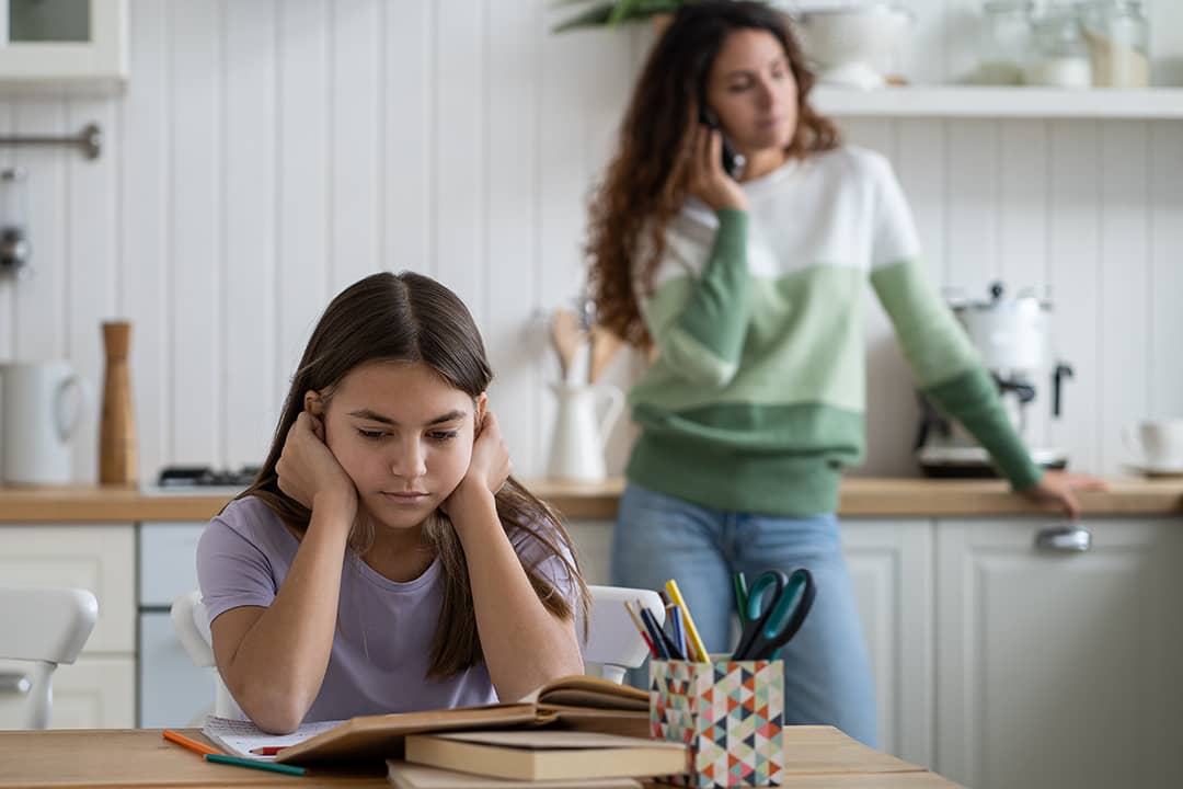 A parent talking to someone over the phone, with her child also struggling to do homework in the dining area.